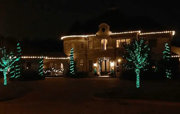 Stone two-story home with warm white roofline lights and trees wrapped in green lights, some spiral wrapped, at night