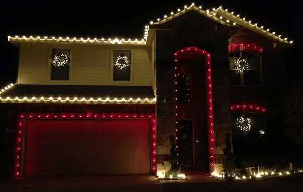 Two-story home with warm white roofline lights, red lights around the garage door and entry arch, and wreaths in windows