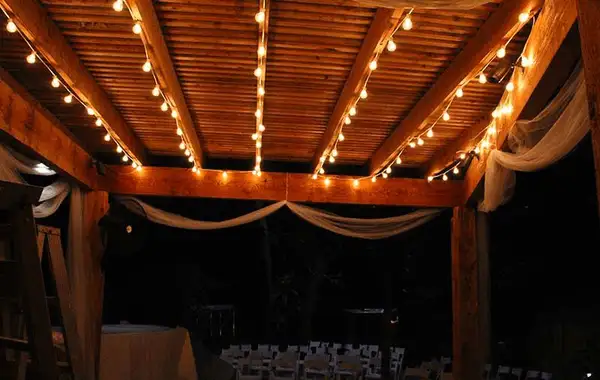 Wood-beamed patio cover with strings of warm white globe lights following the beams and sheer fabric draped at the edges
