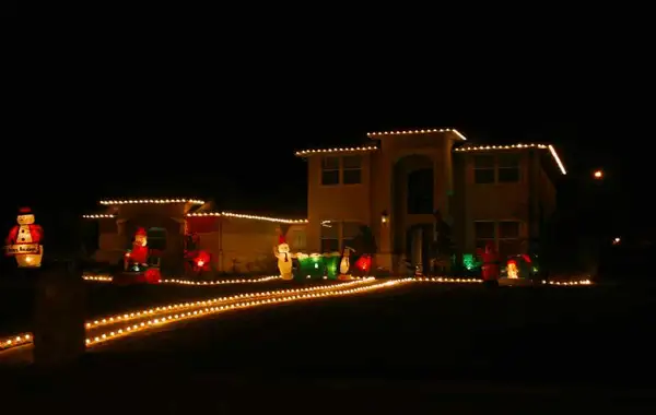 Two-story stucco home at night with warm white roofline lights, a lit driveway border and inflatable holiday figures