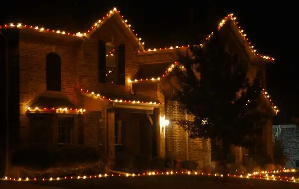 Brick two-story home with red and warm white roofline lights and a lit string along the front lawn at night