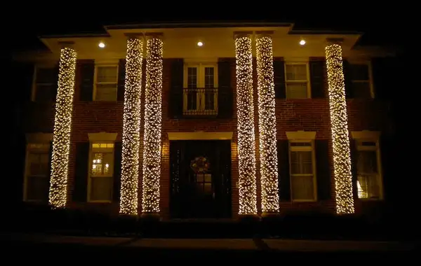 Two-story brick home with columns wrapped in warm white lights and a wreath on the front door at night