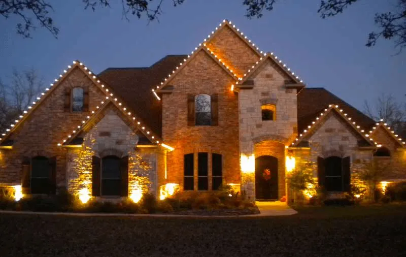 A brick home outlined in warm holiday lighting at dusk