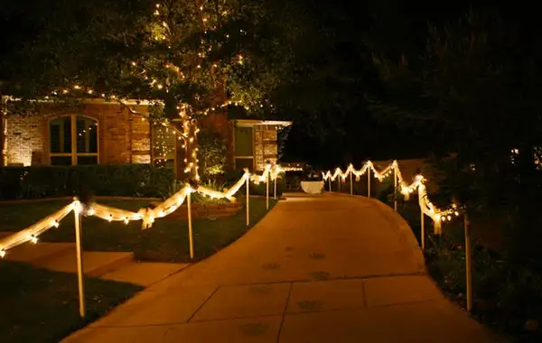 Driveway lined with posts strung with lights and tulle bows, with a light-wrapped tree and brick home beyond at night