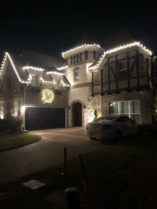 Brick and stone home with warm white lights along rooflines and dormers and a lit wreath above the garage