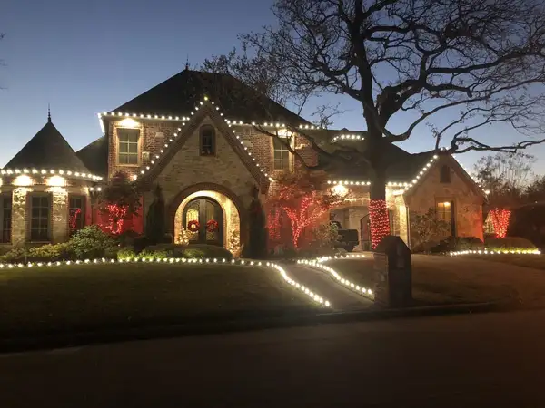Brick and stone two-story home at dusk with warm white roofline lights, red-lit trees, red wreaths and lit walkway borders