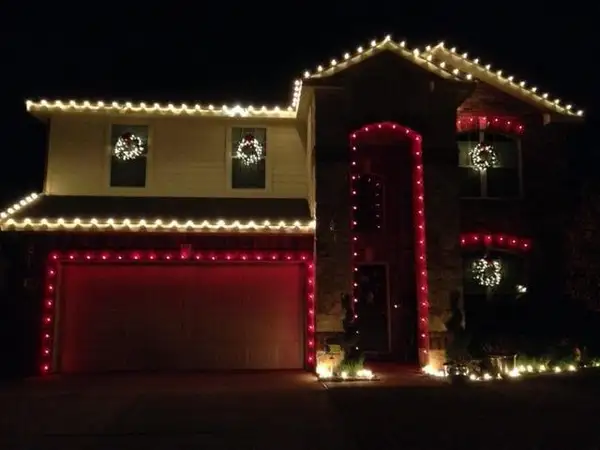 Stone and siding home at night with white eave lights, red-outlined garage door and entry, and lit window wreaths