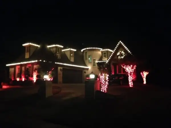 Home at night with warm white lights on dormers, turret and gable, and red-lit trees beside the driveway