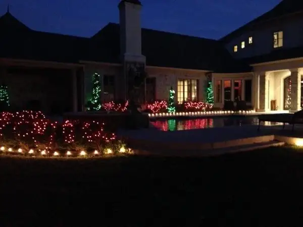 Stone home backyard with red-lit shrubs, green-lit cone-shaped trees and warm white lights along the pool edge at dusk