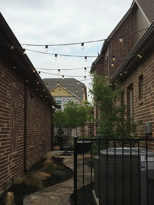 Narrow side yard between brick homes with festoon bulb string lights crossing overhead above a flagstone path