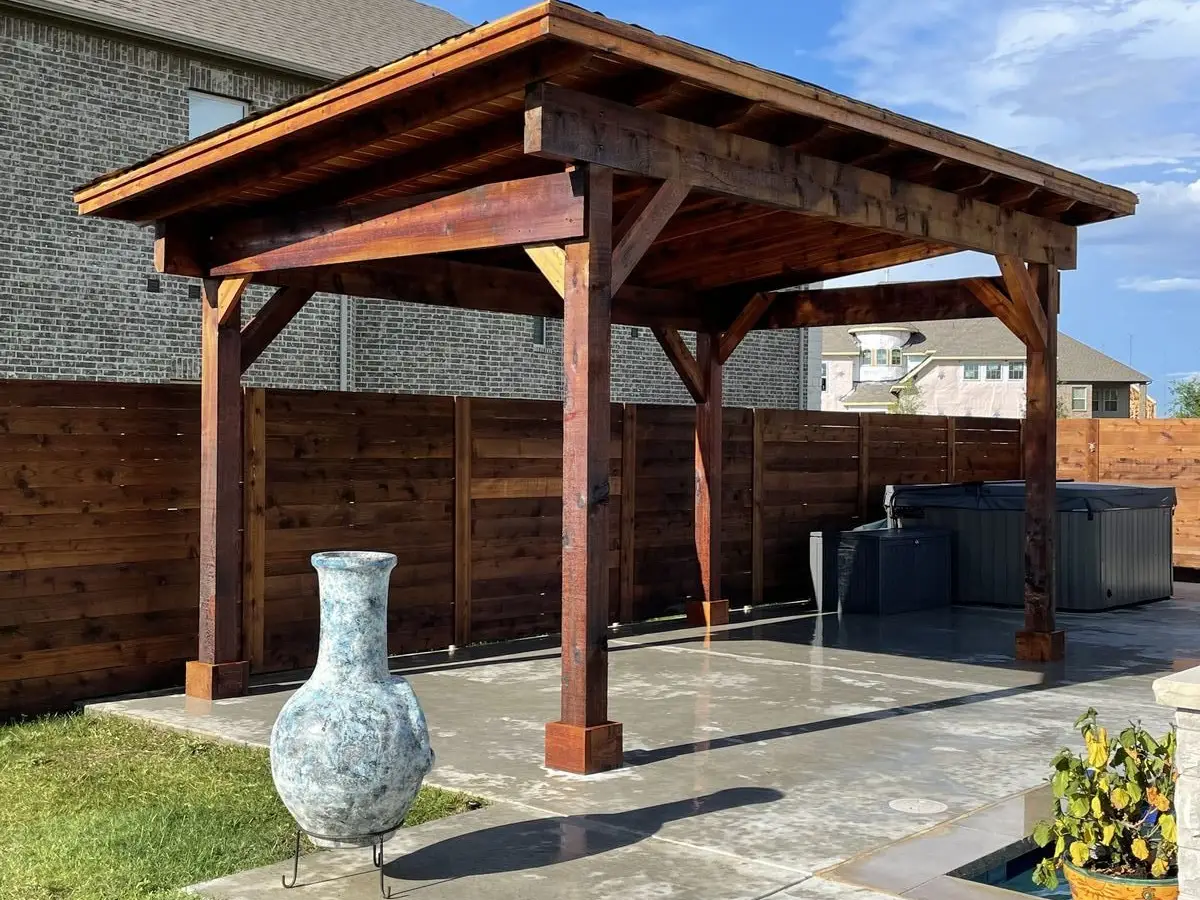 Freestanding stained cedar pavilion with heavy posts and knee braces over a concrete patio beside a hot tub