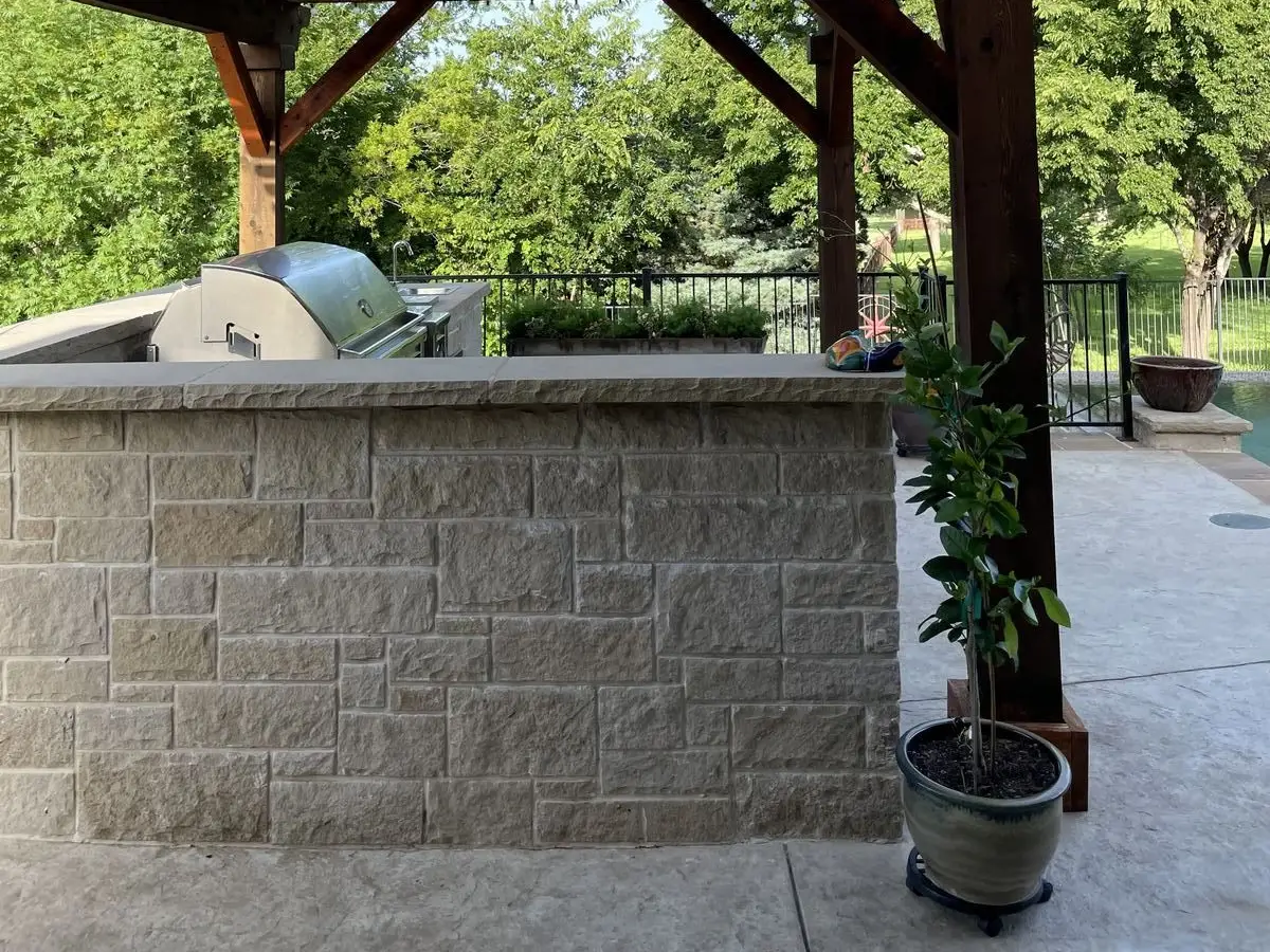 Cedar louvered pergola over a stone outdoor kitchen island with a built-in grill beside a pool