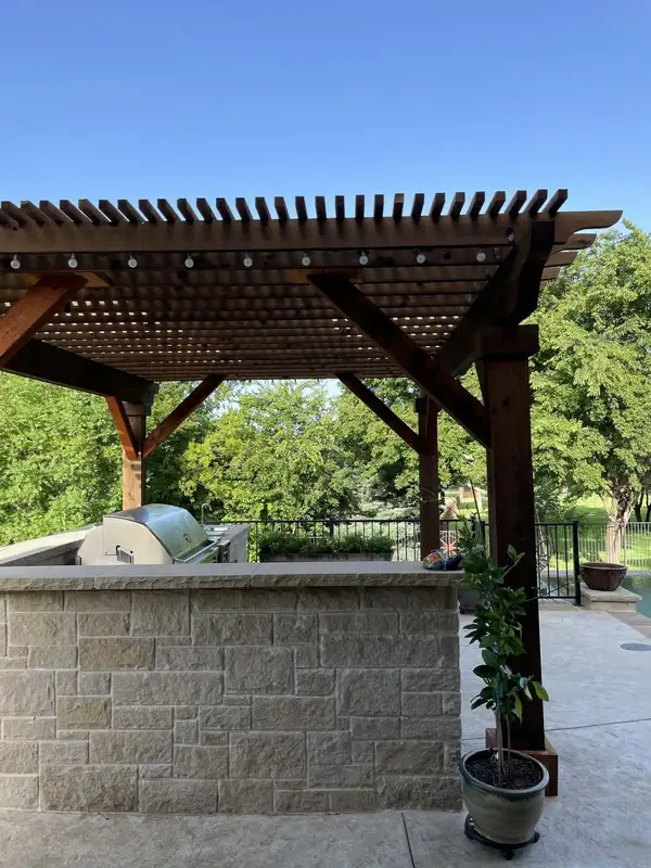 Cedar louvered pergola over a stone outdoor kitchen island with a built-in grill beside a pool