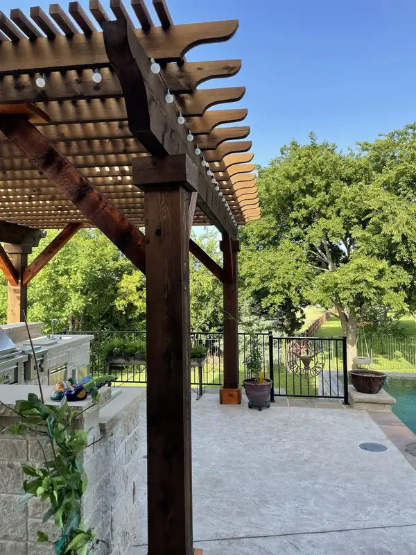 Cedar pergola post with scrolled rafter ends and string lights beside an outdoor kitchen and pool