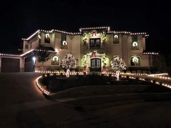 Stucco home with warm white roofline lights, wreaths with red bows, entry garland and two fully lit trees at night