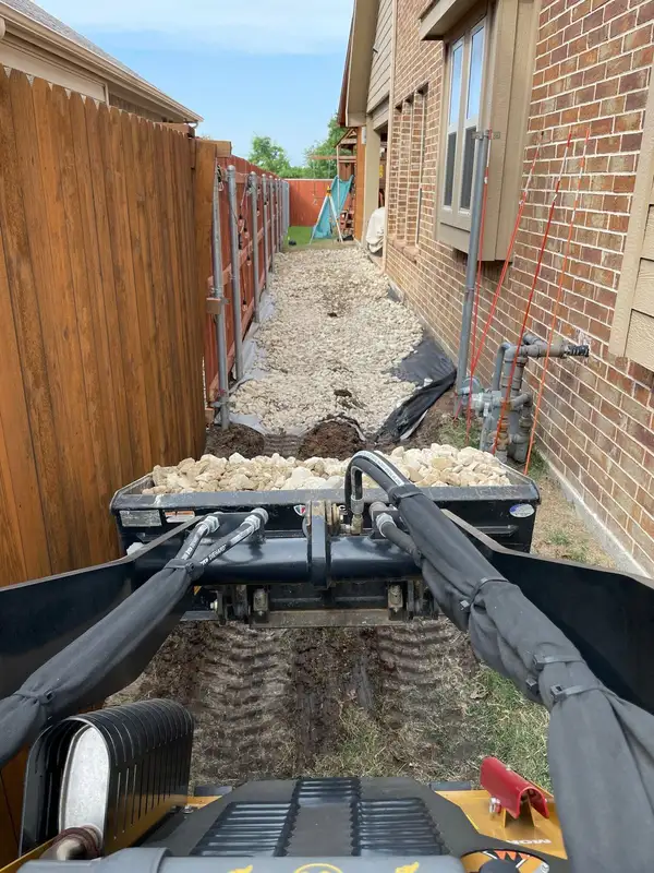 Mini skid steer bucket of white rock facing a side yard trench lined with fabric and corrugated pipe