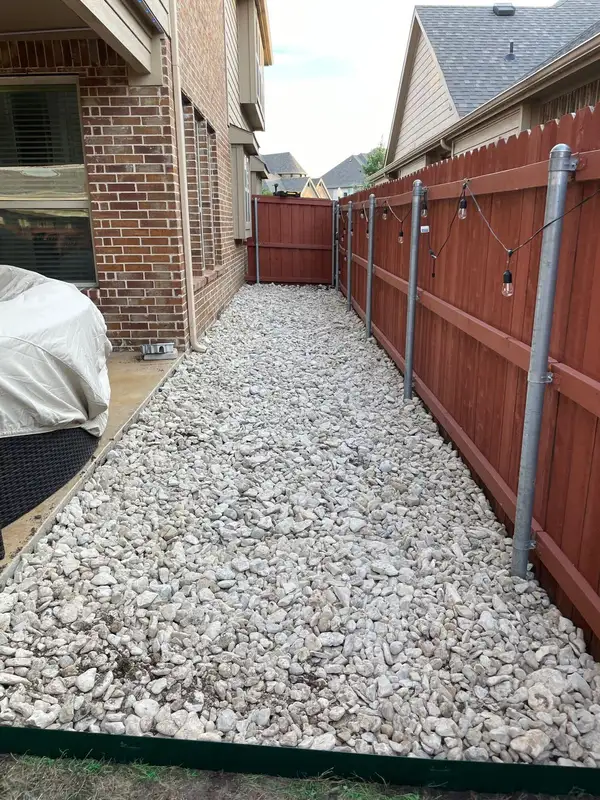 Narrow side yard filled with white crushed rock between a brick house and a red-stained fence with string lights