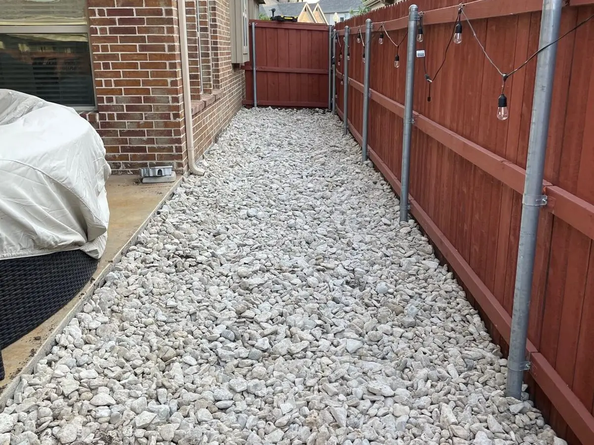 Narrow side yard filled with white crushed rock between a brick house and a red-stained fence with string lights