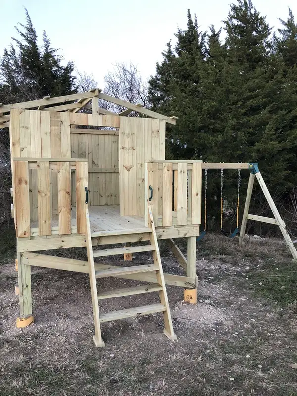 Unstained pressure-treated wood playhouse on stilts with a ladder and swing set under construction