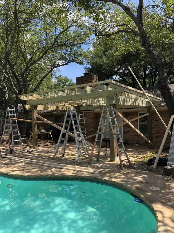 Pressure-treated pergola framing under construction beside a backyard pool, with ladders and lumber on the patio