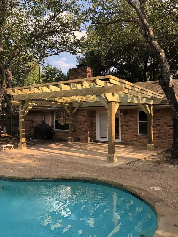 Pressure-treated wood pergola with open rafters and angled brackets over a stamped concrete patio beside a pool