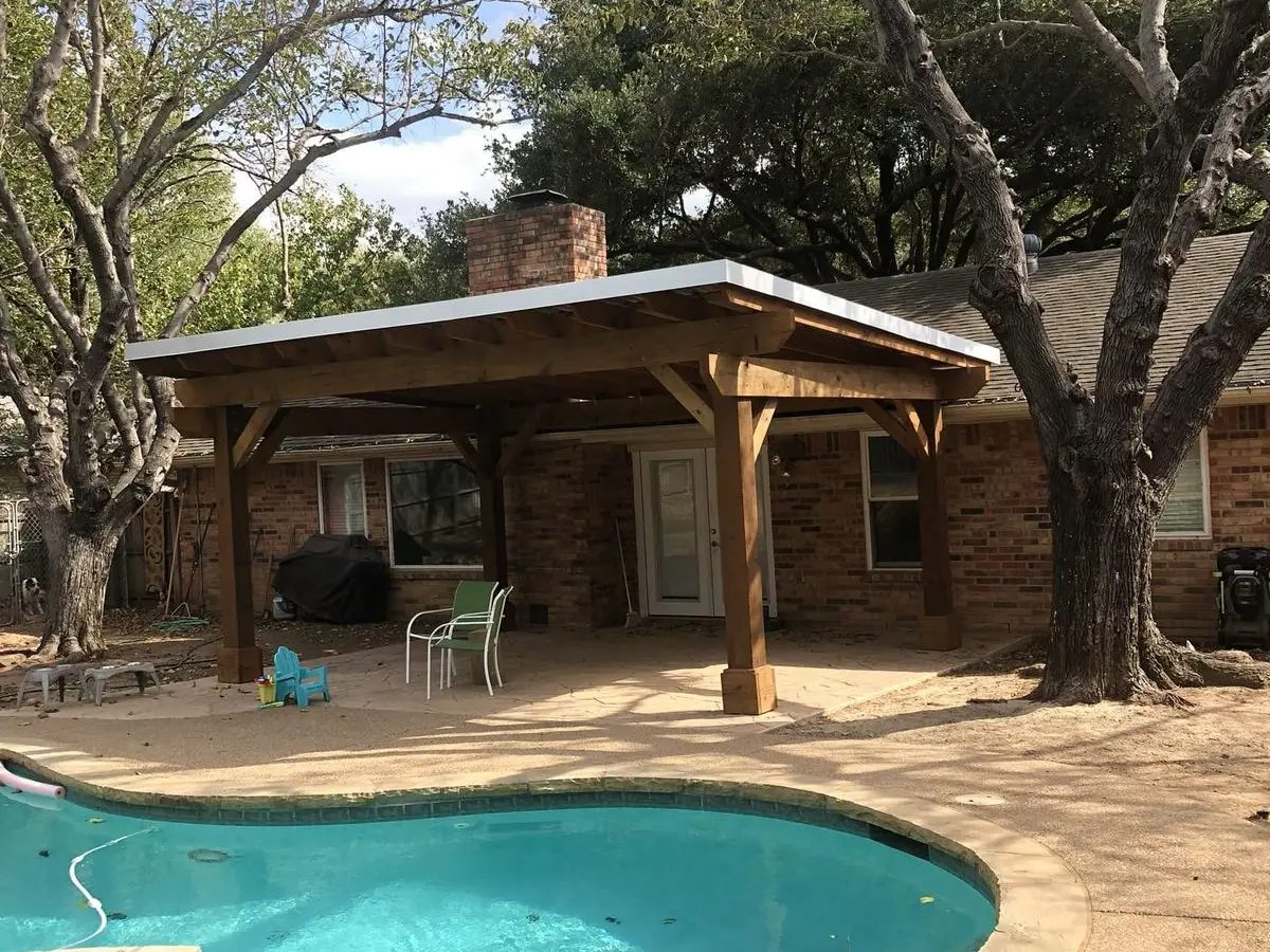 Stained wood patio cover with a low-slope white roof over a stamped concrete patio beside a pool