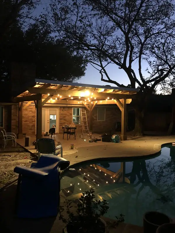Wood patio cover at dusk with string lights overhead, beside a pool behind a brick home