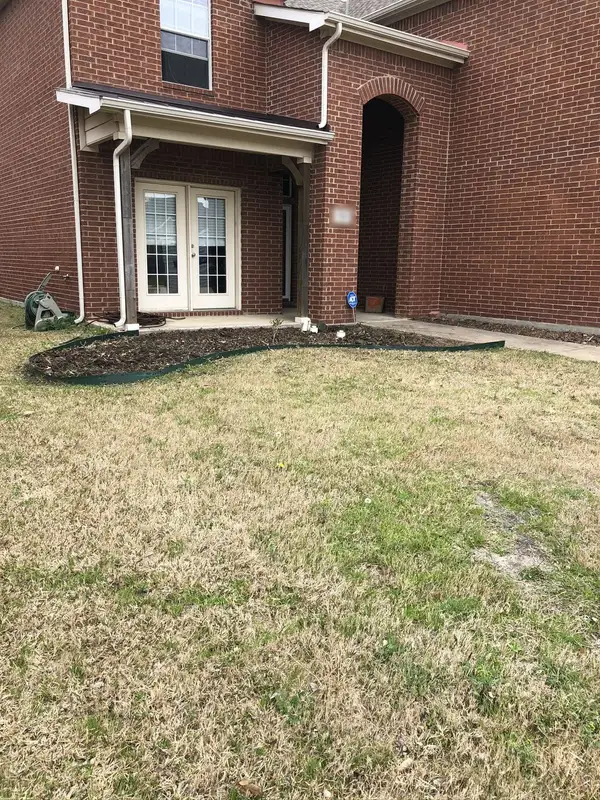 Curved steel-edged mulch bed beside a brick home's porch with a patchy dormant lawn