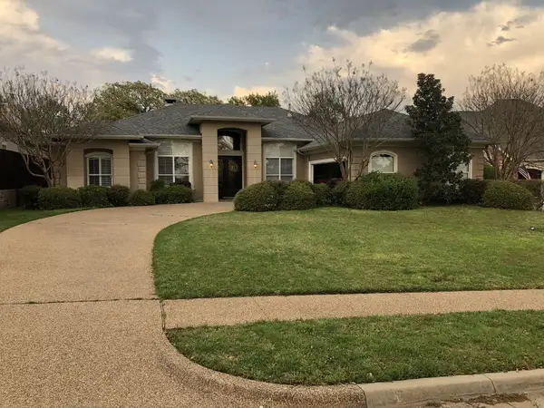 Single-story stucco home with a curved driveway, trimmed shrubs and bare crape myrtles under a cloudy dusk sky