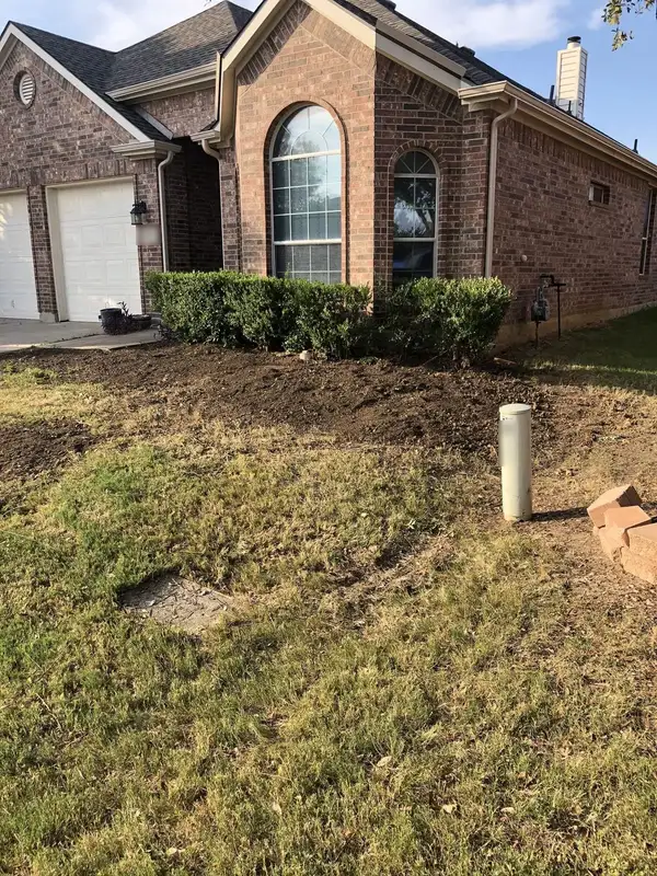 Freshly graded soil bed in front of trimmed shrubs on a patchy dry front lawn at a brick home