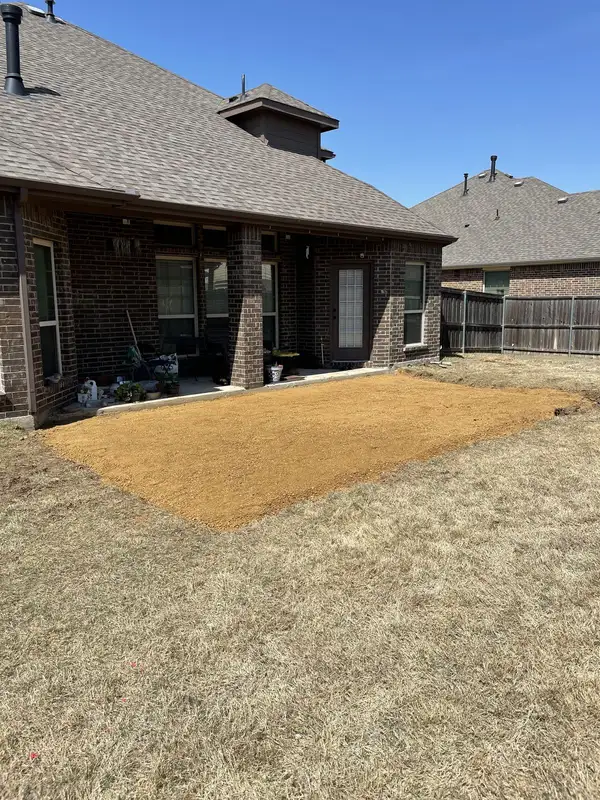 Leveled orange crushed-rock base against a covered patio slab on a dark brick home with a dormant lawn